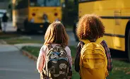 Two children stand together, backpacks on, facing a line of yellow school buses on a sunny day.